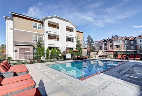 A swimming pool is surrounded by red lounge chairs in front of apartment buildings.