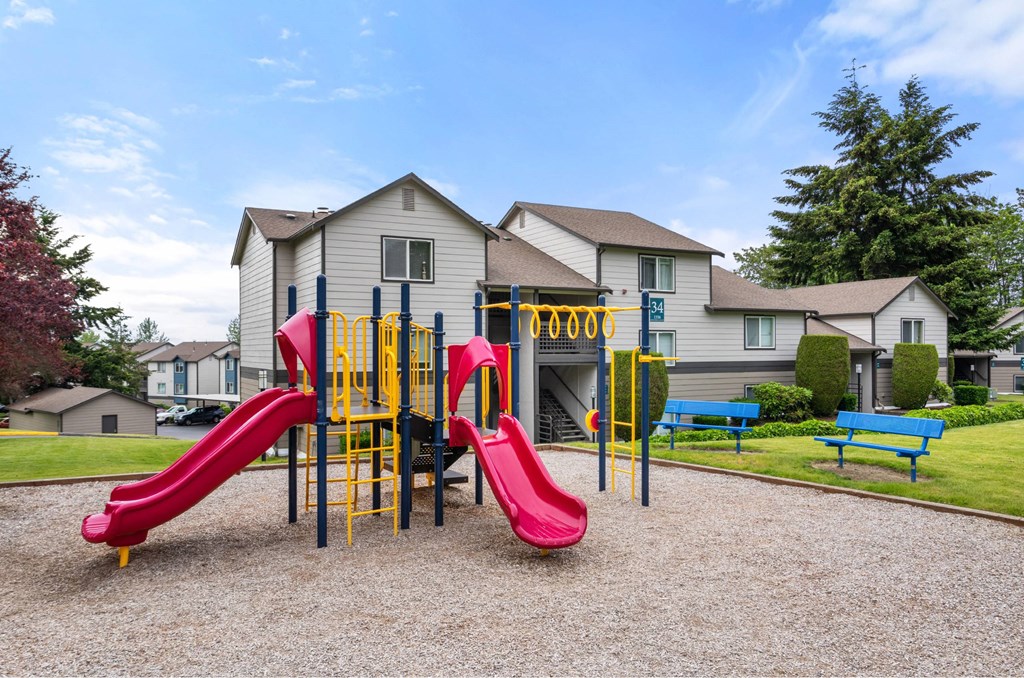 A playground with a red slide and yellow and blue climbing structures in front of a building.