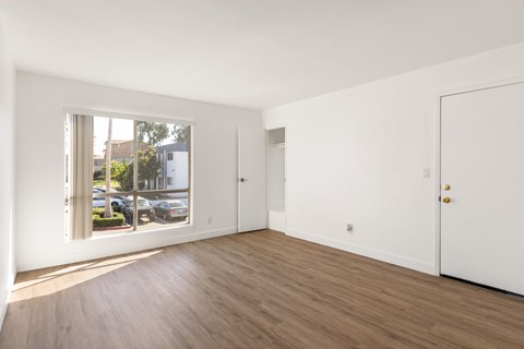 A room with white walls and wooden flooring, with a window showing a parking lot outside.