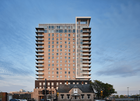 A tall building with a mix of brick and glass facade stands in front of a smaller house and a tree.