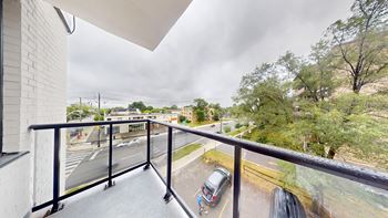 A balcony with a metal railing overlooks a parking lot and a tree-lined street.