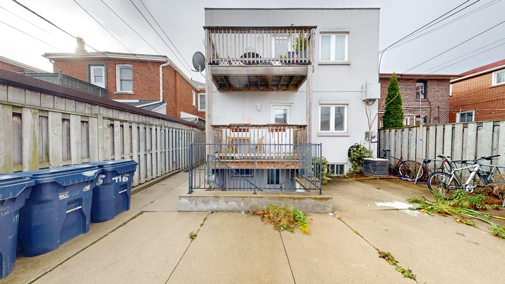A grey building with a balcony is in the middle of a courtyard with blue bins and a fence.