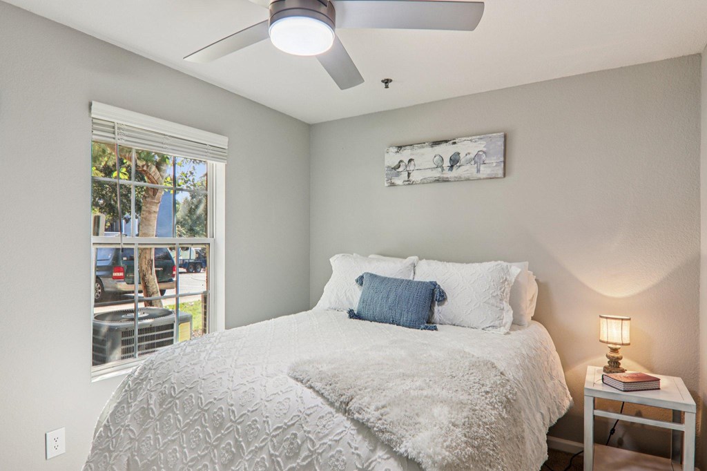 A bedroom with a bed, a ceiling fan, and a window with a view of a parking lot.