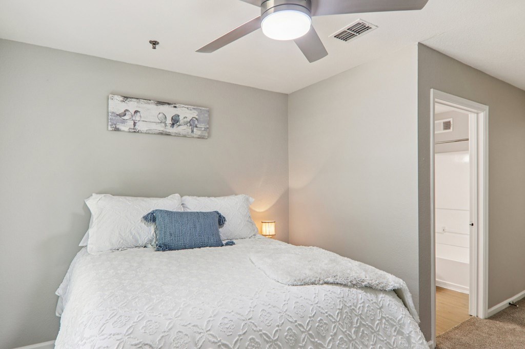 A bedroom with a bed, pillows, a ceiling fan, and a painting on the wall.