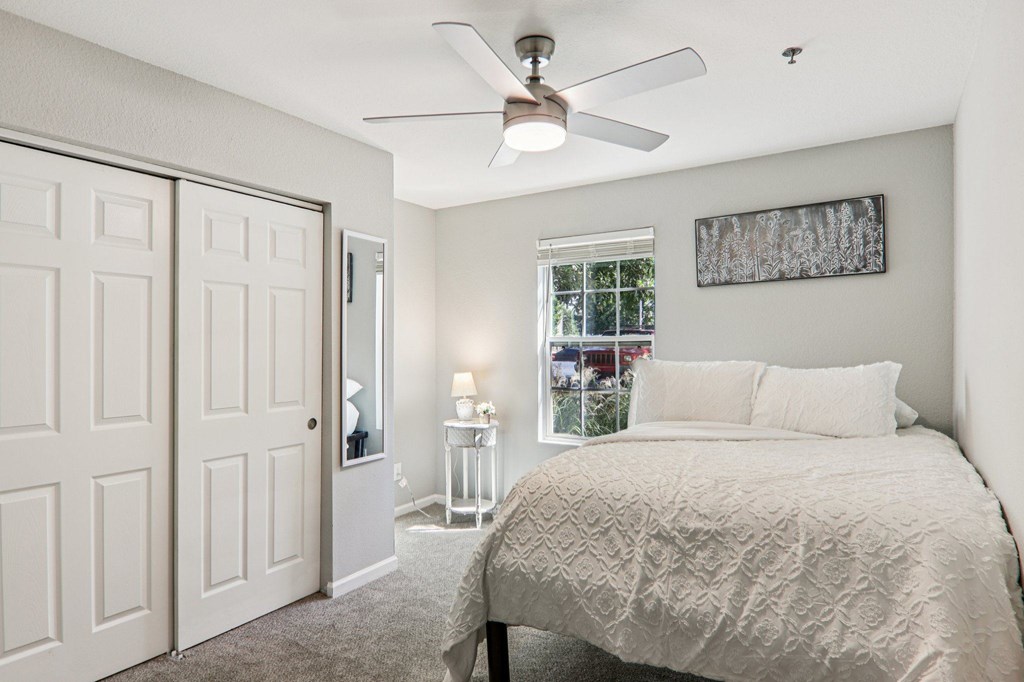 A bedroom with a bed, a ceiling fan, and a window with a view of the city.