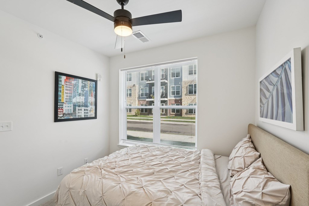 A bedroom with a bed, a ceiling fan, and a window overlooking a street.