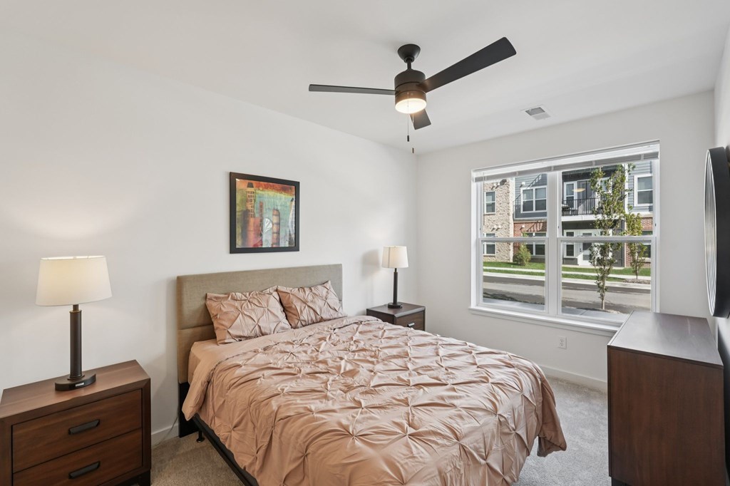 A bedroom with a bed, two lamps, a ceiling fan, and a window overlooking a street.