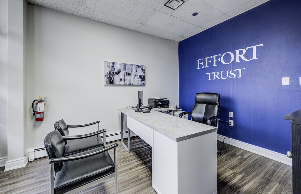 A room with a white desk and a black chair with a blue wall that says Effort Trust.
