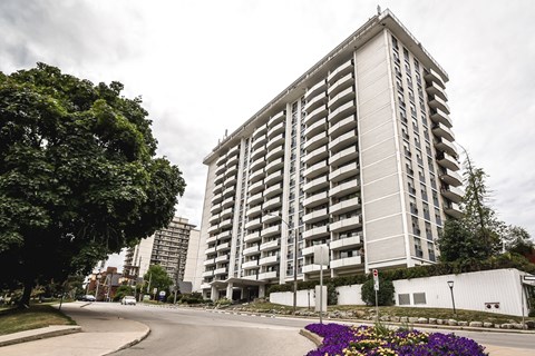 A tall residential building with balconies is surrounded by trees and a street with a flower bed.