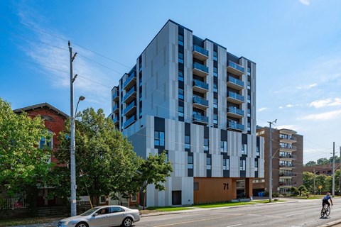 A modern multi-story building with blue and white balconies is situated on a street corner with a car and a cyclist in the foreground.