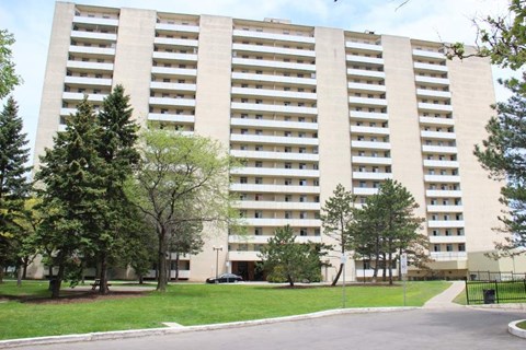 A large beige building with many windows and balconies, surrounded by trees and a paved walkway.
