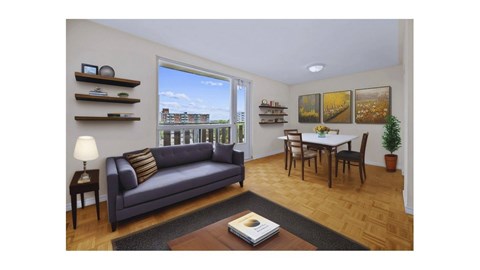 A living room with a grey couch, a small table with a lamp, a dining table with chairs, and pictures on the wall.