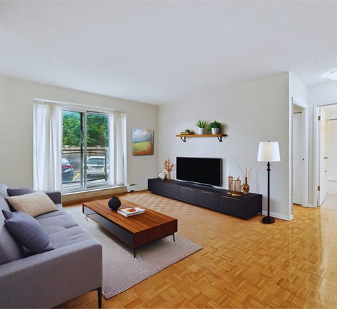 A living room with a grey couch, a wooden coffee table, a black TV stand, and a large window.