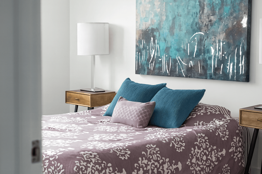 A bed with a purple comforter and two blue pillows sits in a room with a painting on the wall.