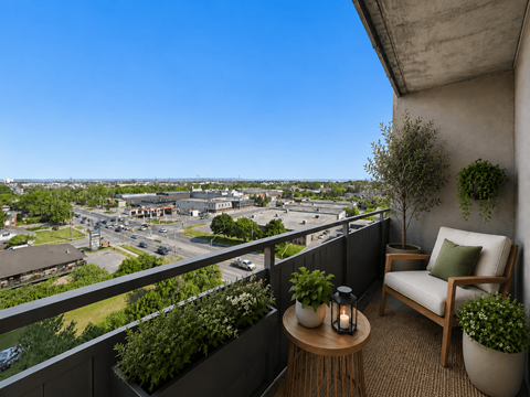 A balcony with a chair, table, and potted plants overlooks a city street.