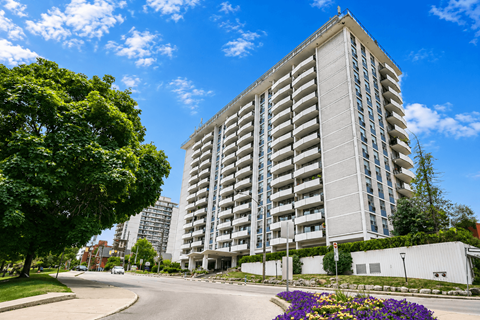 A tall residential building with balconies is surrounded by greenery and a clear blue sky.