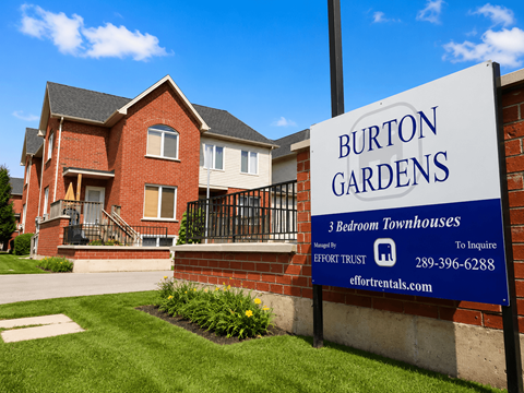 A sign for Burton Gardens 3 bedroom townhouses stands in front of a brick building.