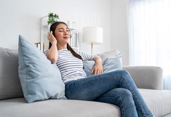 A woman is sitting on a grey couch with a blue pillow, wearing a striped shirt and blue jeans, and appears to be listening to music with headphones.at The Maverick, Ontario, N2H 0E3