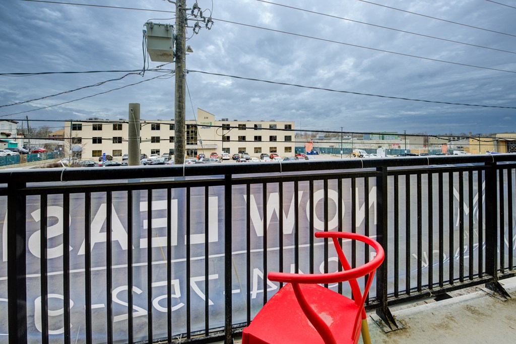 A red chair sits on a balcony with a view of a parking lot and buildings.