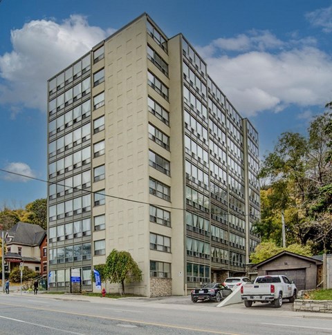 A tall beige building with many windows is surrounded by trees and parked cars.