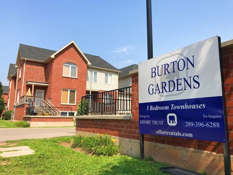 A sign for Burton Gardens 3 Bedroom Townhouses stands in front of a brick house.