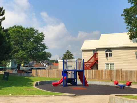A playground with a blue and red slide in the foreground and a house with a red staircase in the background.