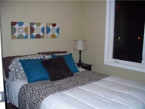 A bed with a white comforter and a blue pillow is in front of a wall with three colorful paintings.