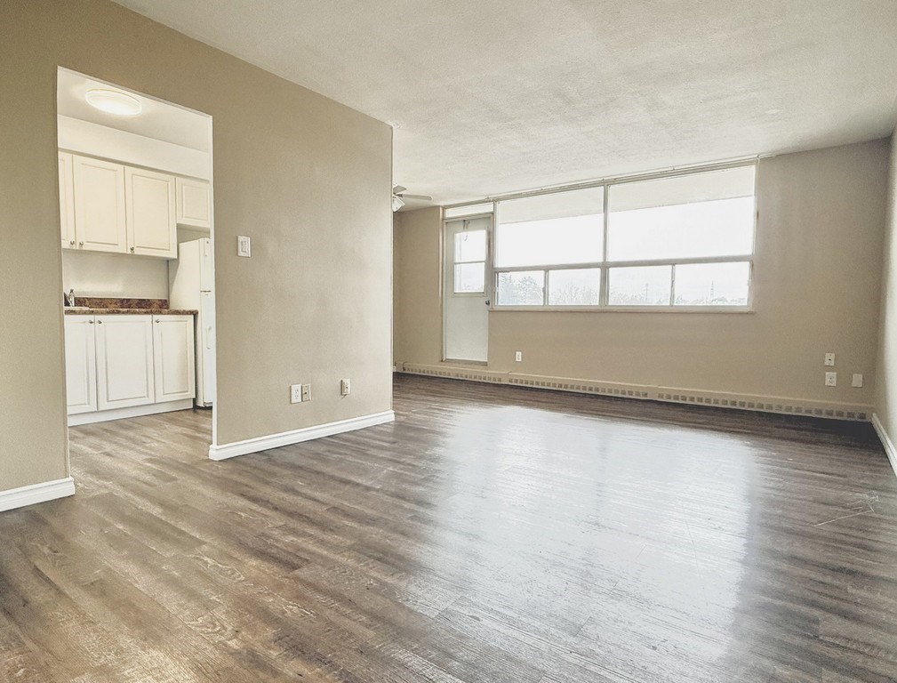 A large empty room with wooden flooring and a kitchen area in the background.