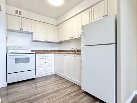 A white refrigerator stands in a kitchen with wooden floors and white cabinets.