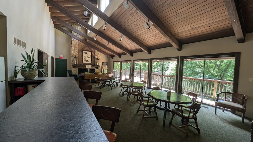A room with a bar area, tables with chairs, and a balcony with a view of trees.