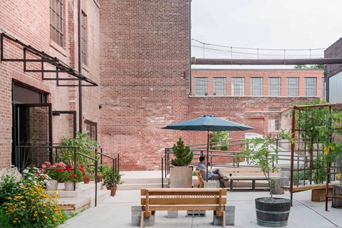 A man is sitting on a bench under a blue umbrella in a courtyard with plants and flowers.