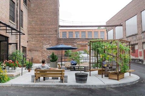 A person is sitting on a bench under a blue umbrella in a courtyard with plants and a trellis.