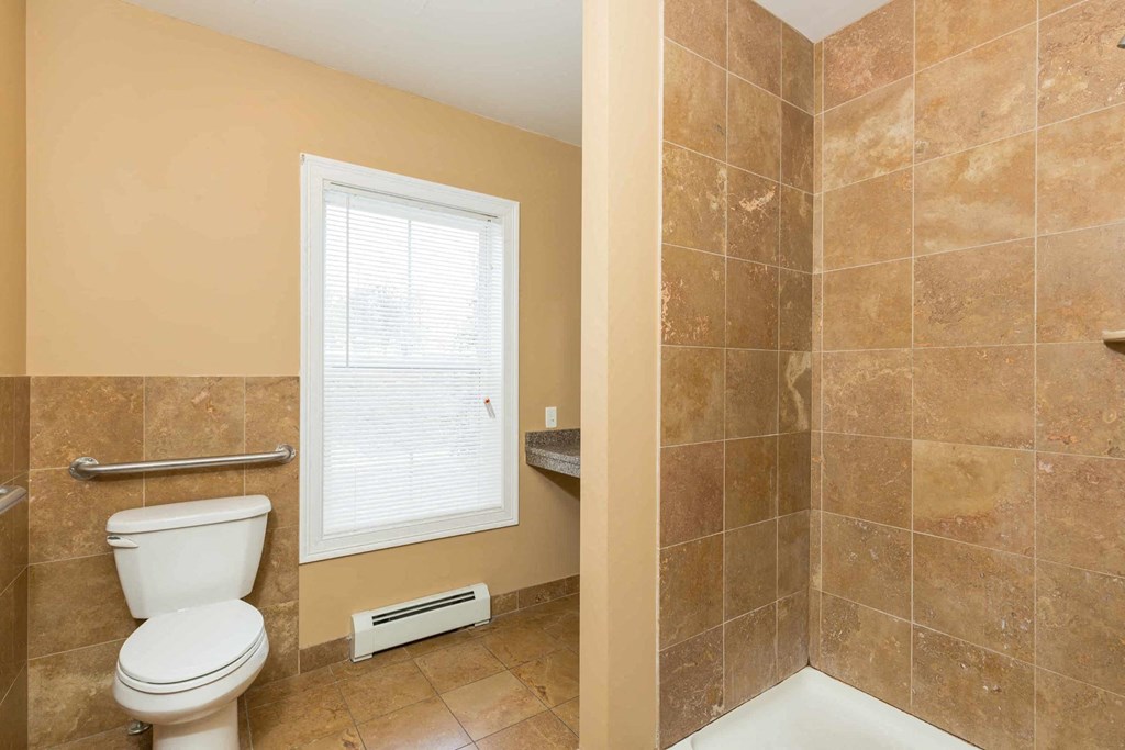A bathroom with a toilet, tiled shower, and window.