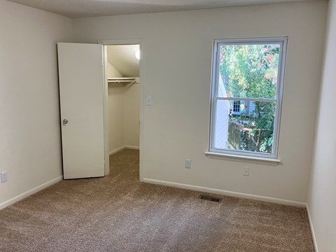 A room with a carpeted floor, a white door, and a window with a view of trees and a house outside.