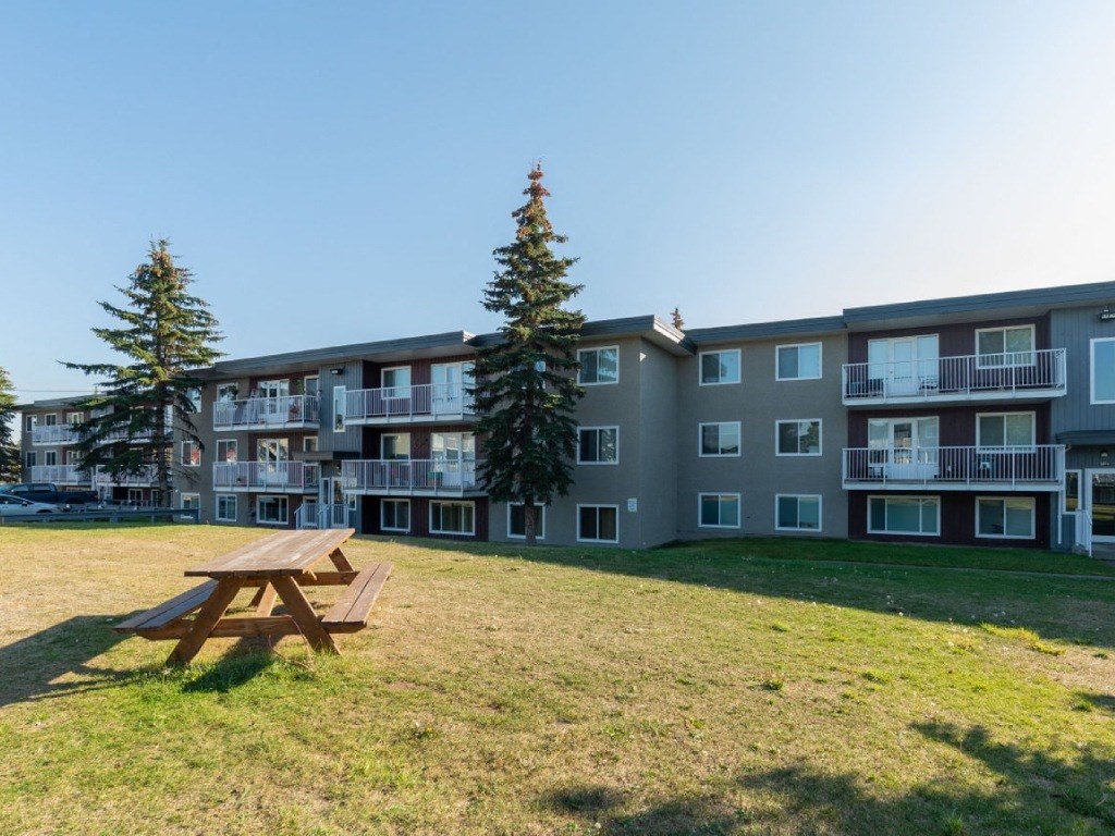 A wooden picnic table is in the foreground of a grassy area in front of apartment buildings.