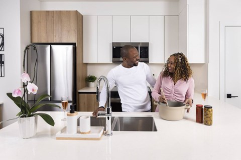 A man and woman are standing in a kitchen with a sink and a refrigerator.