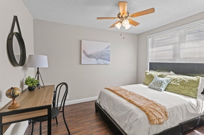 A bedroom with a bed, a table, a chair, a ceiling fan, and a painting on the wall.