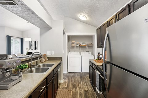 A modern kitchen with a stainless steel refrigerator, a washer and dryer stacked in the corner, and a dining area with a table and chairs.
