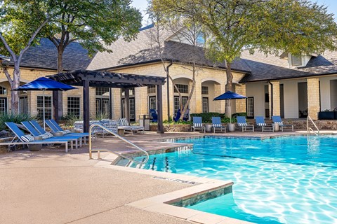 A pool area with a building in the background and lounge chairs and umbrellas.