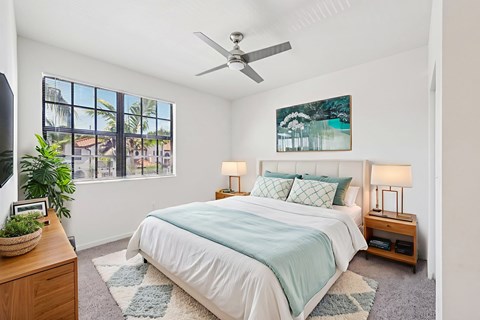 A bedroom with a bed, a ceiling fan, and a window with a view of a cityscape.