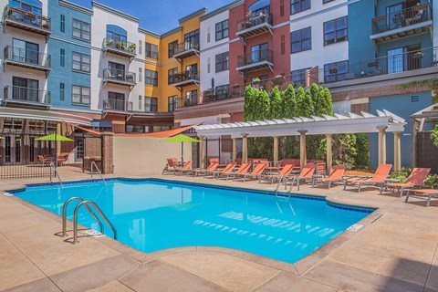A pool surrounded by chairs and umbrellas in front of apartment buildings.