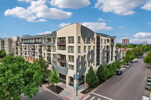 A modern building with a curved design is surrounded by trees and a street with cars.