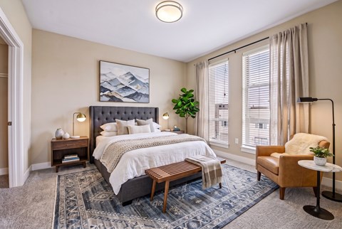 A bedroom with a large bed, a bench, a chair, and a plant.