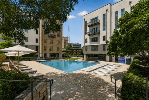 A pool surrounded by a patio and trees with apartment buildings in the background.