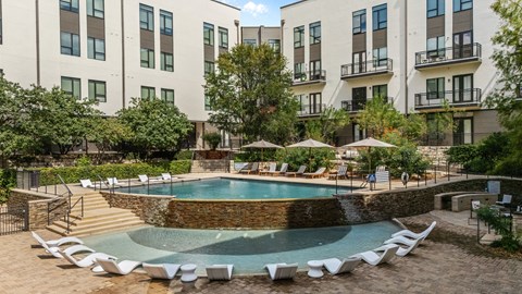 A swimming pool surrounded by chairs and umbrellas in front of apartment buildings.