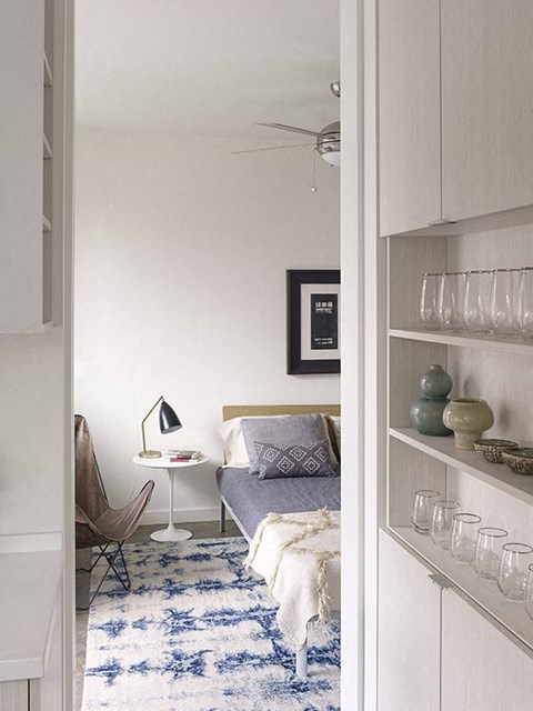 A bedroom with a bed, a chair, a nightstand with a lamp, and a shelf with glasses on it.