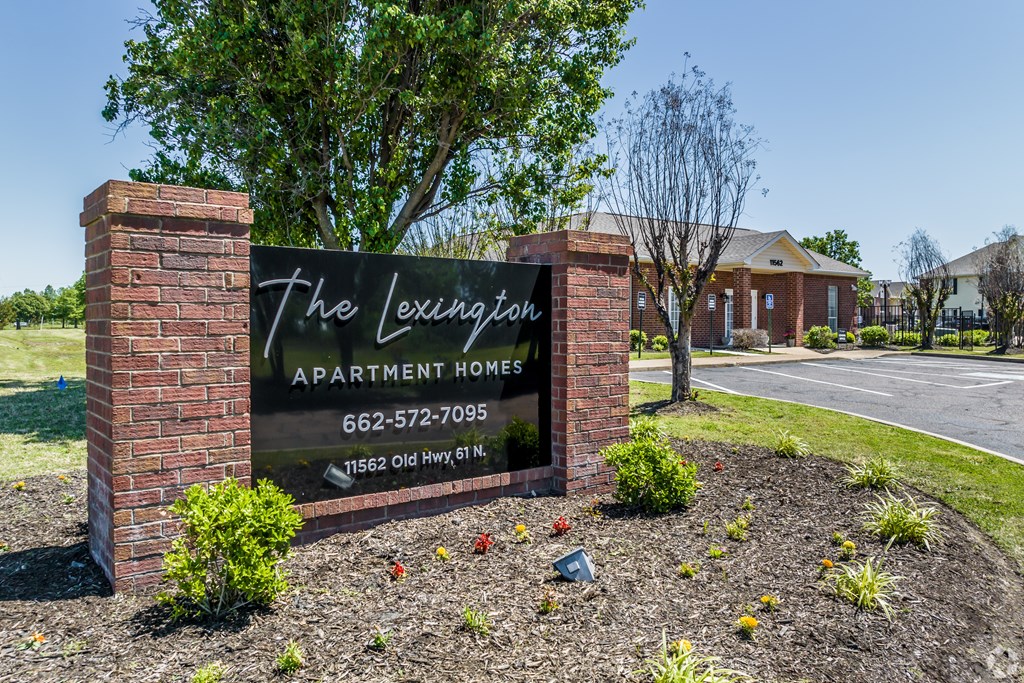 A sign for The Lexington Apartment Homes stands in front of a tree and a building.