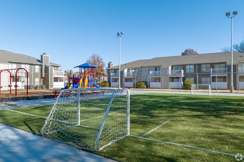 A soccer goal is in the foreground of a grassy area with apartment buildings in the background.
