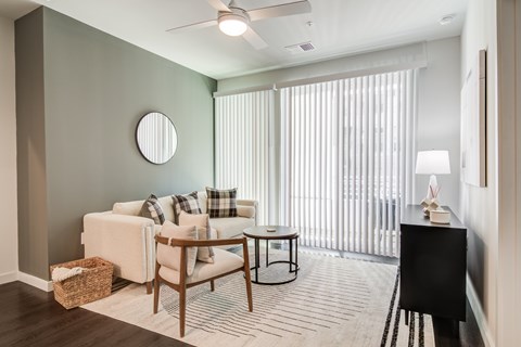 A living room with a white couch, a wooden chair, a round mirror, a small table, a lamp, and a window with blinds.