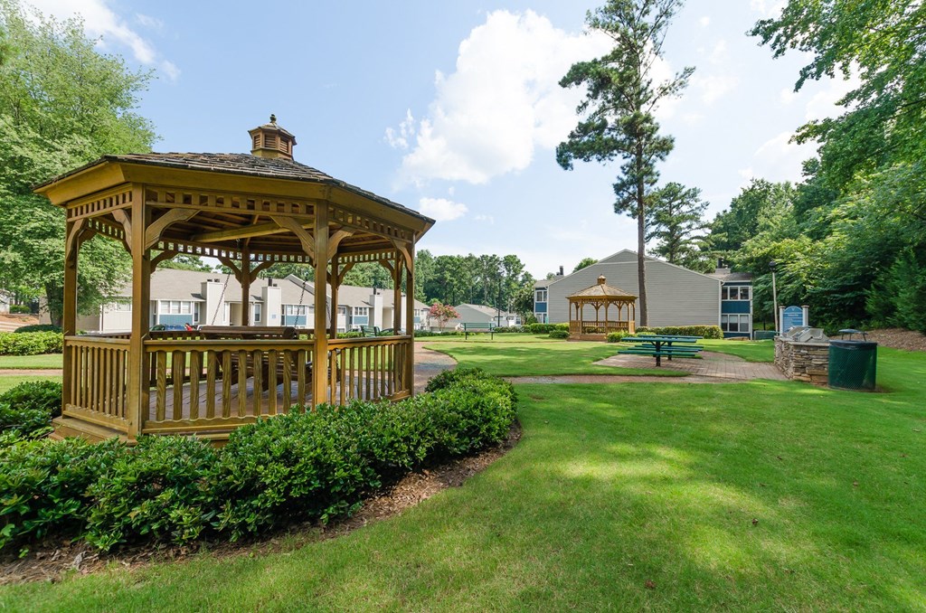 A wooden gazebo is surrounded by green bushes and trees.